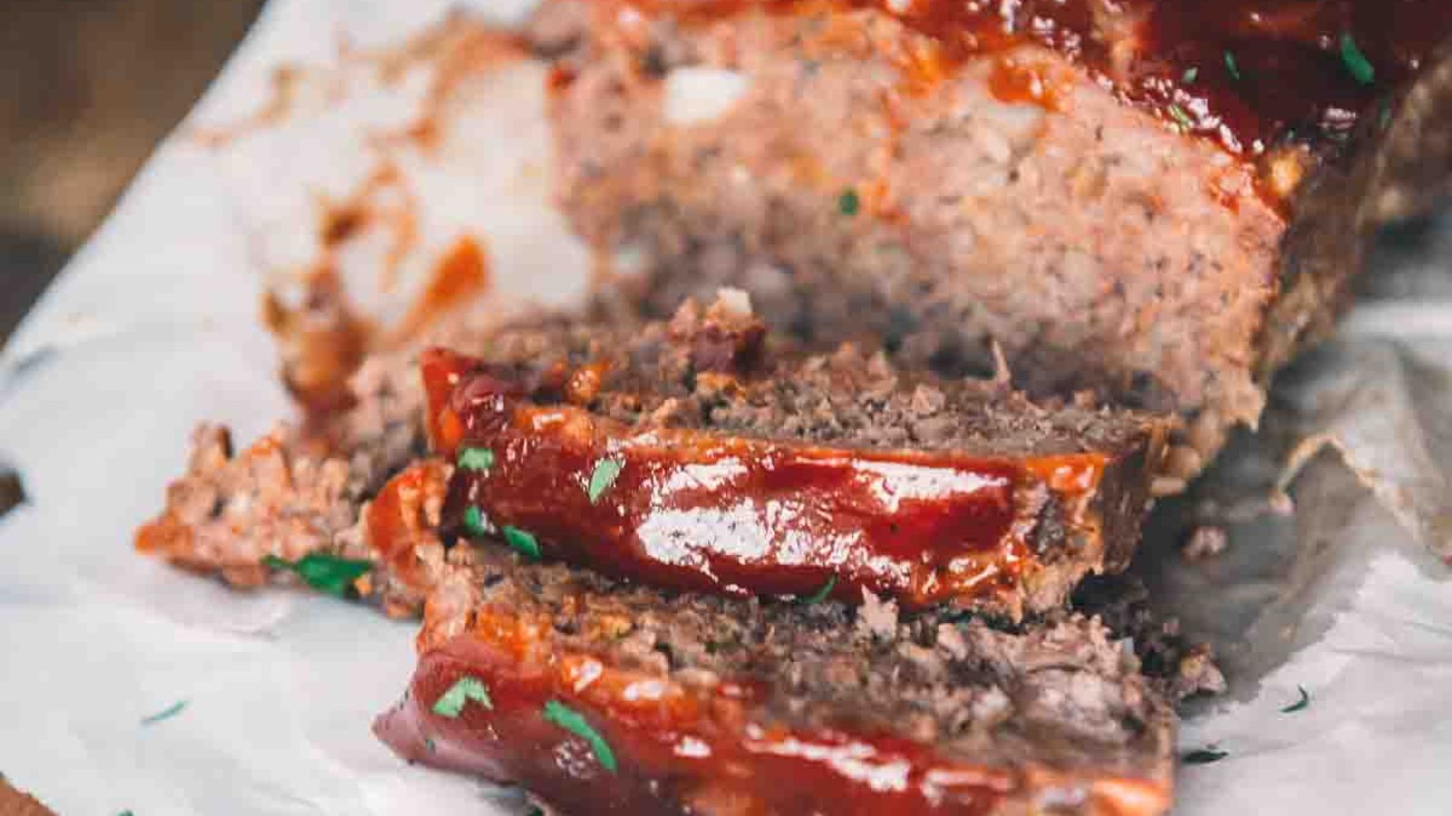 Close-up of sliced meatloaf topped with glaze, garnished with herbs, on parchment paper.