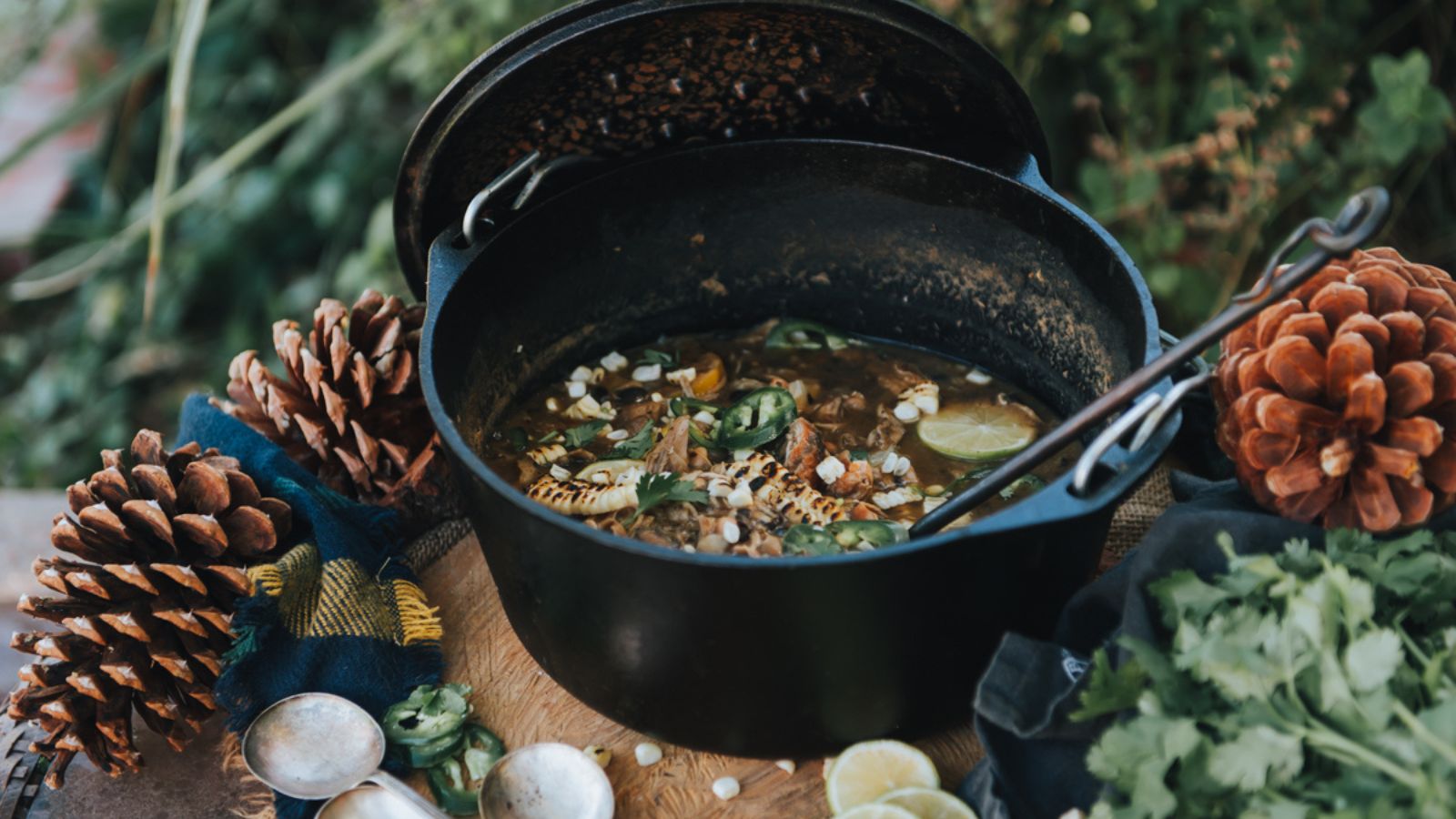 A pot of stew with meat, corn, and jalapeños surrounded by cilantro, pine cones, and lime slices, placed on a wooden surface.