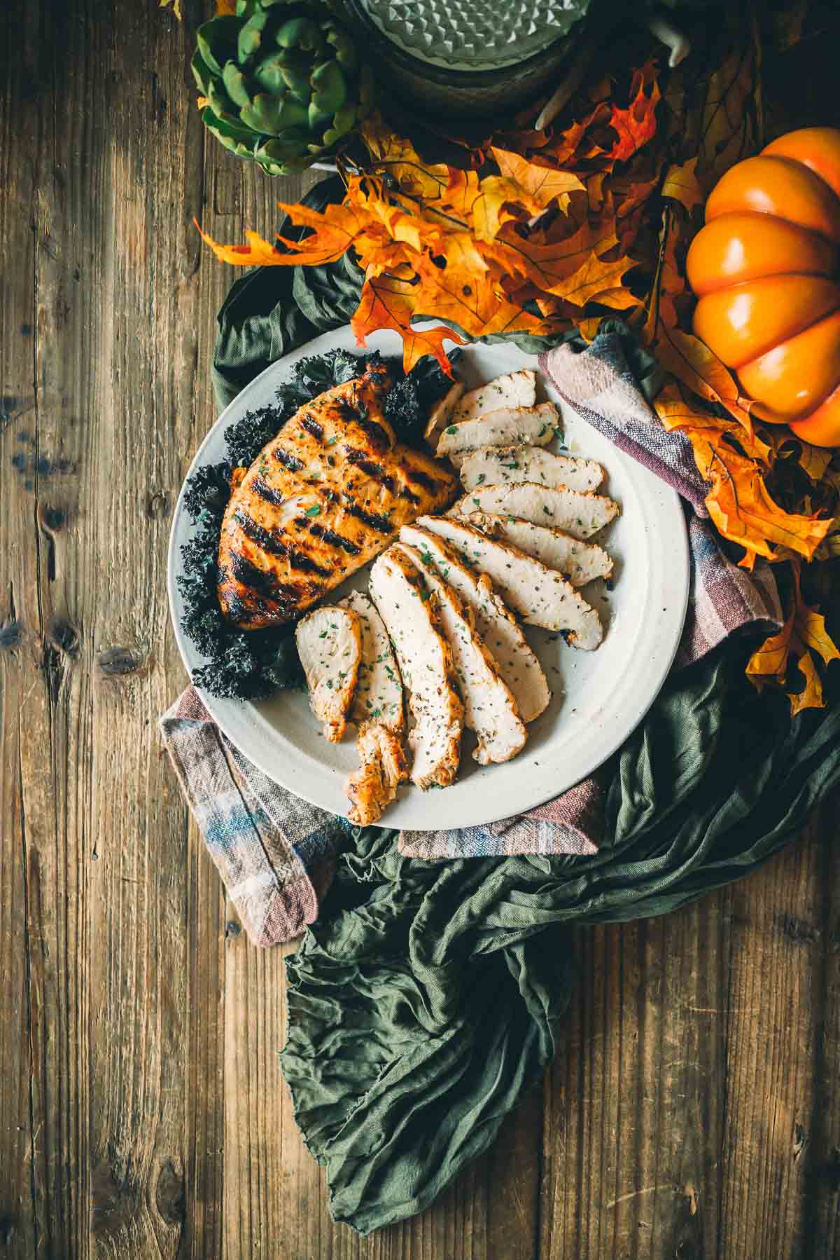 A plate of sliced grilled turkey tenderloin garnished with kale, surrounded by autumn leaves, a pumpkin, and artichokes, on a wooden table.