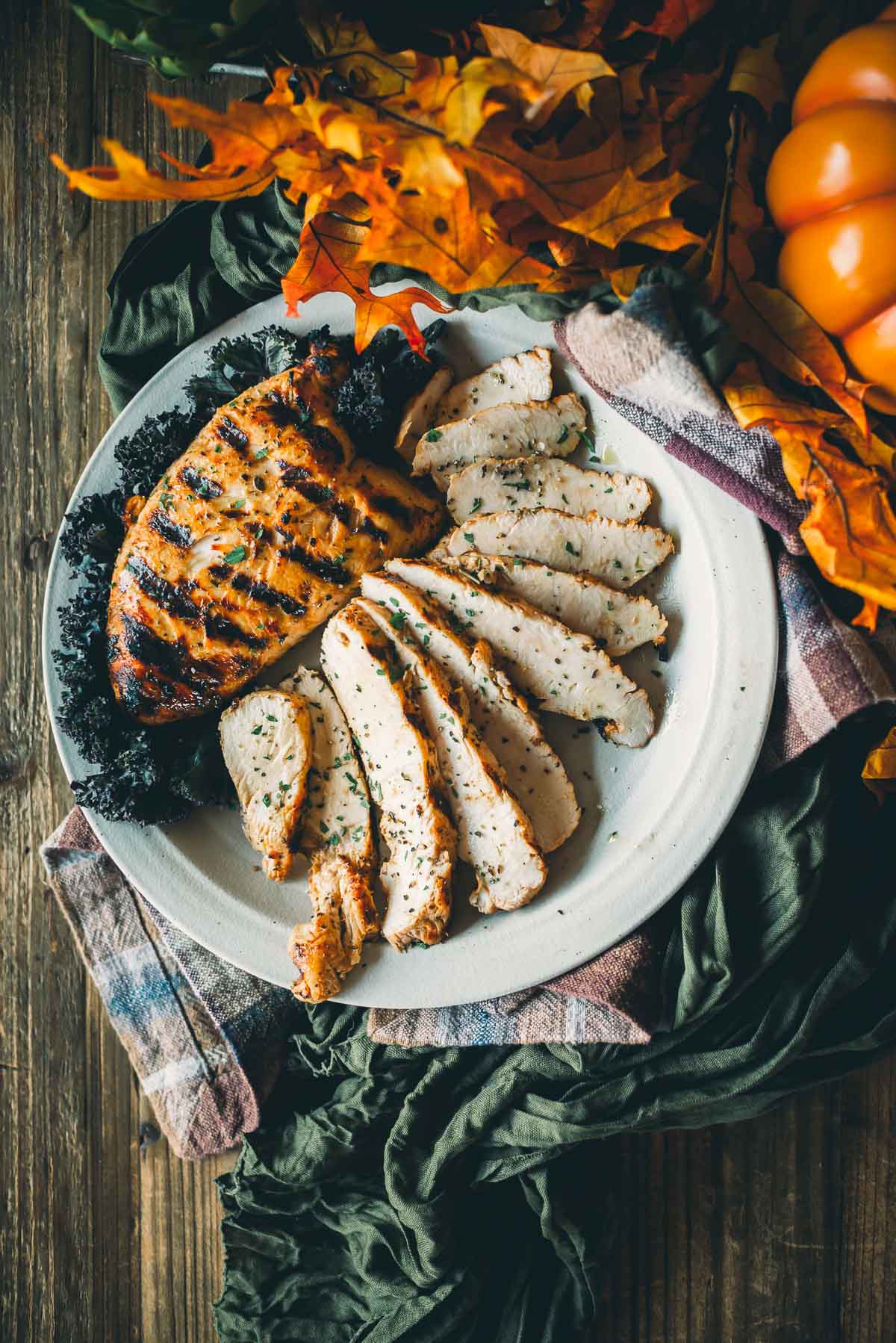 Grilled turkey tenderloin slices on a white plate, garnished with herbs and kale, surrounded by fall leaves and a small pumpkin on a wooden table.