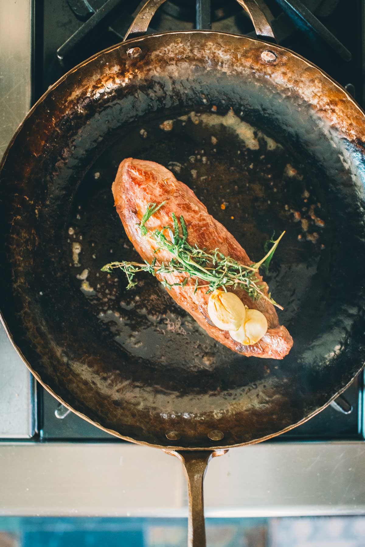 A teres major steak with sprigs of rosemary and slices of garlic is cooking in a copper pan on a stovetop.