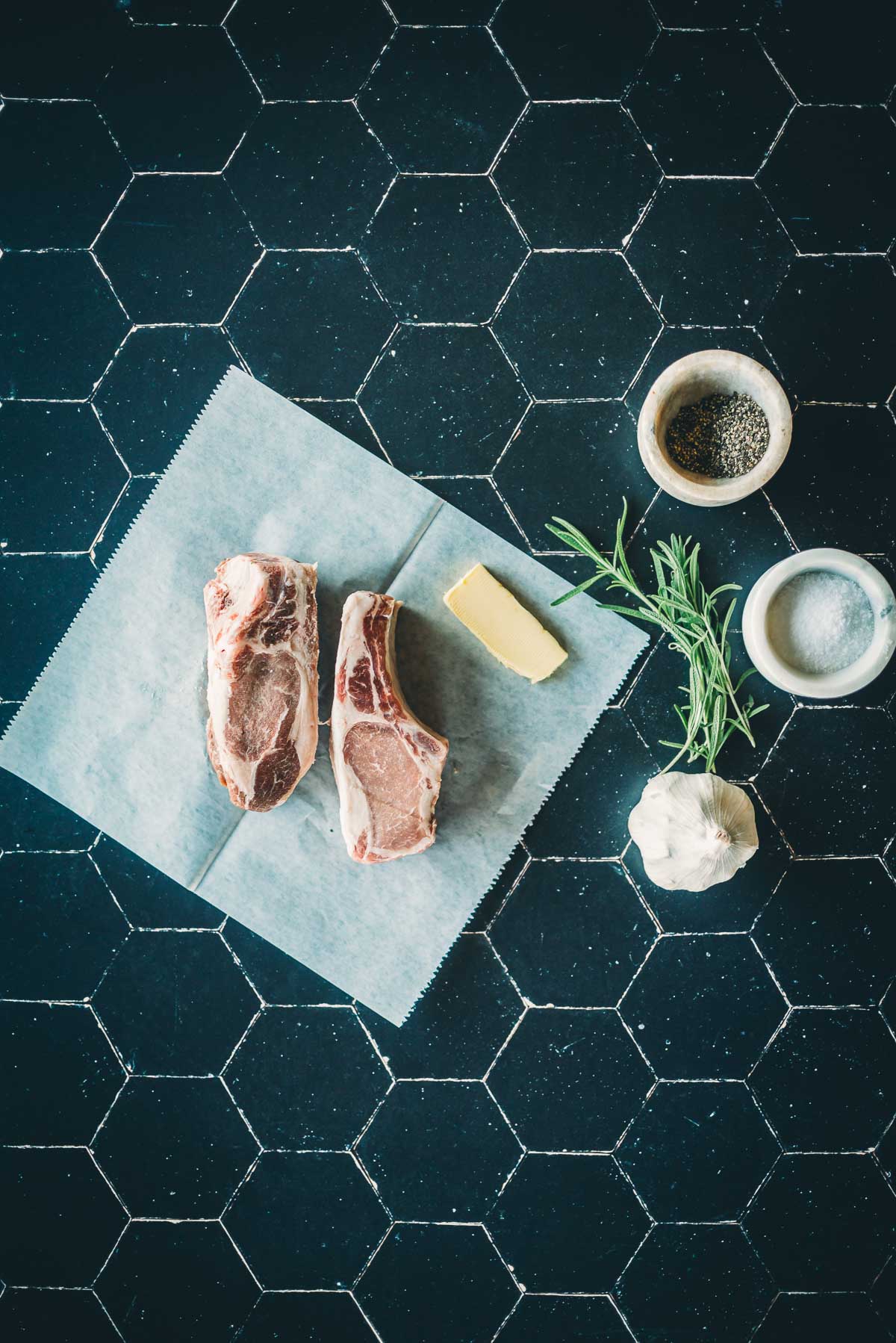 Two raw boar chops on parchment paper, next to a pat of butter, a garlic bulb, rosemary sprig, and bowls of salt and pepper on a black tile surface.