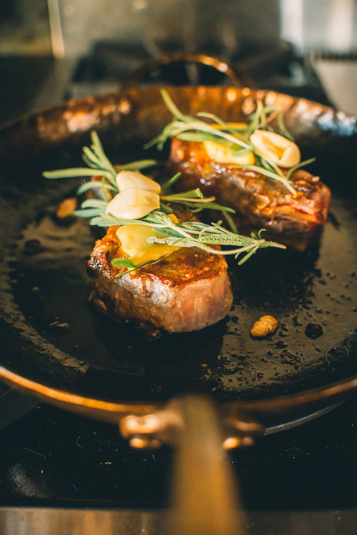 Two boar chops cooking in a pan, topped with rosemary and garlic cloves.