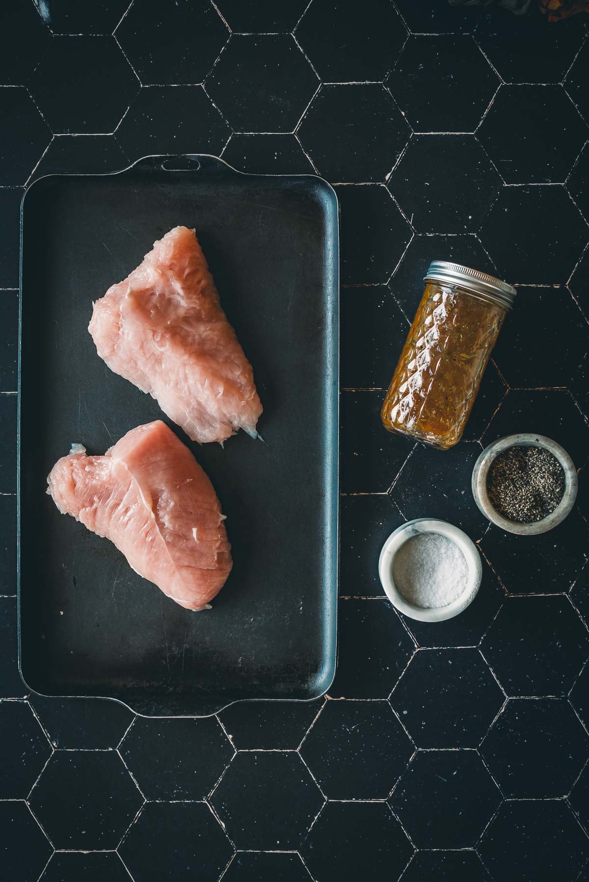 Two raw turkey tenderloin breasts on a tray, accompanied by a jar of sauce and bowls of salt and pepper, placed on a black tile surface.