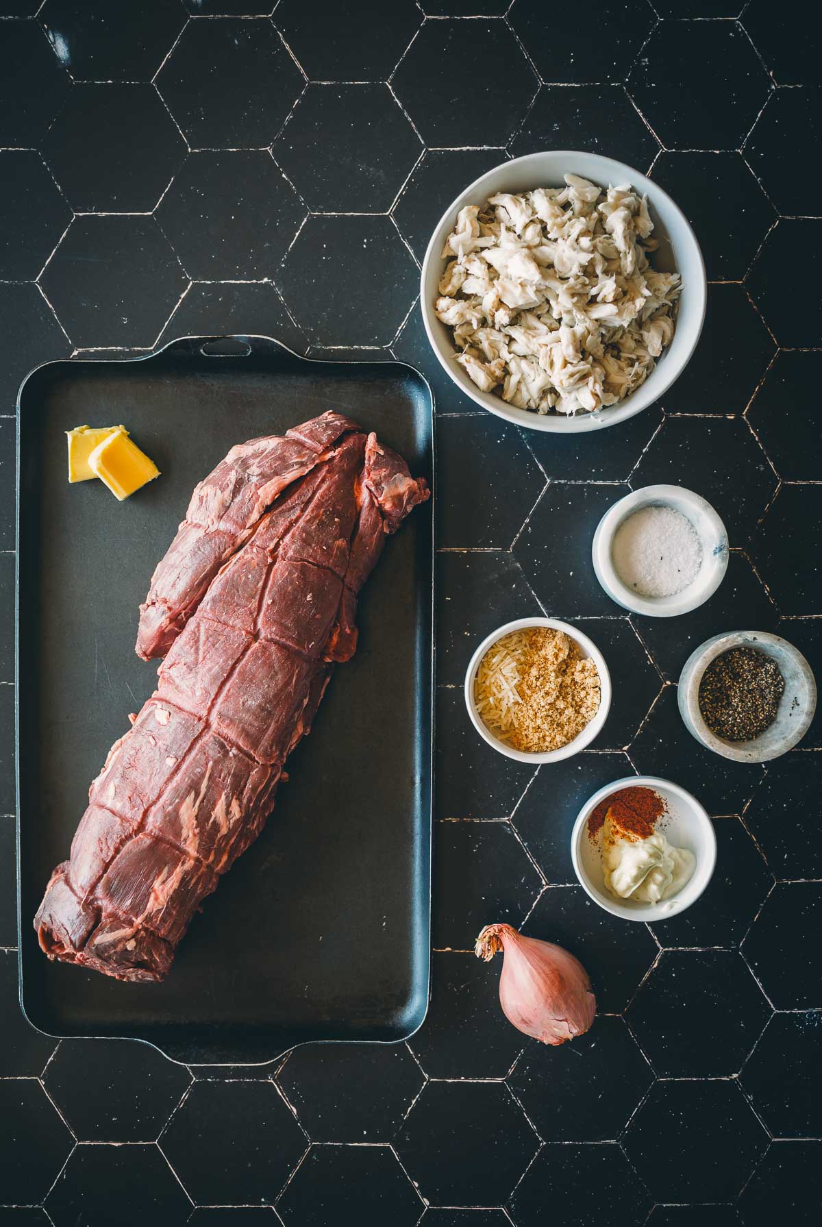 Raw beef tenderloin on a tray with butter, alongside bowls of crab, salt, breadcrumbs, pepper, and spices on a black tile surface.