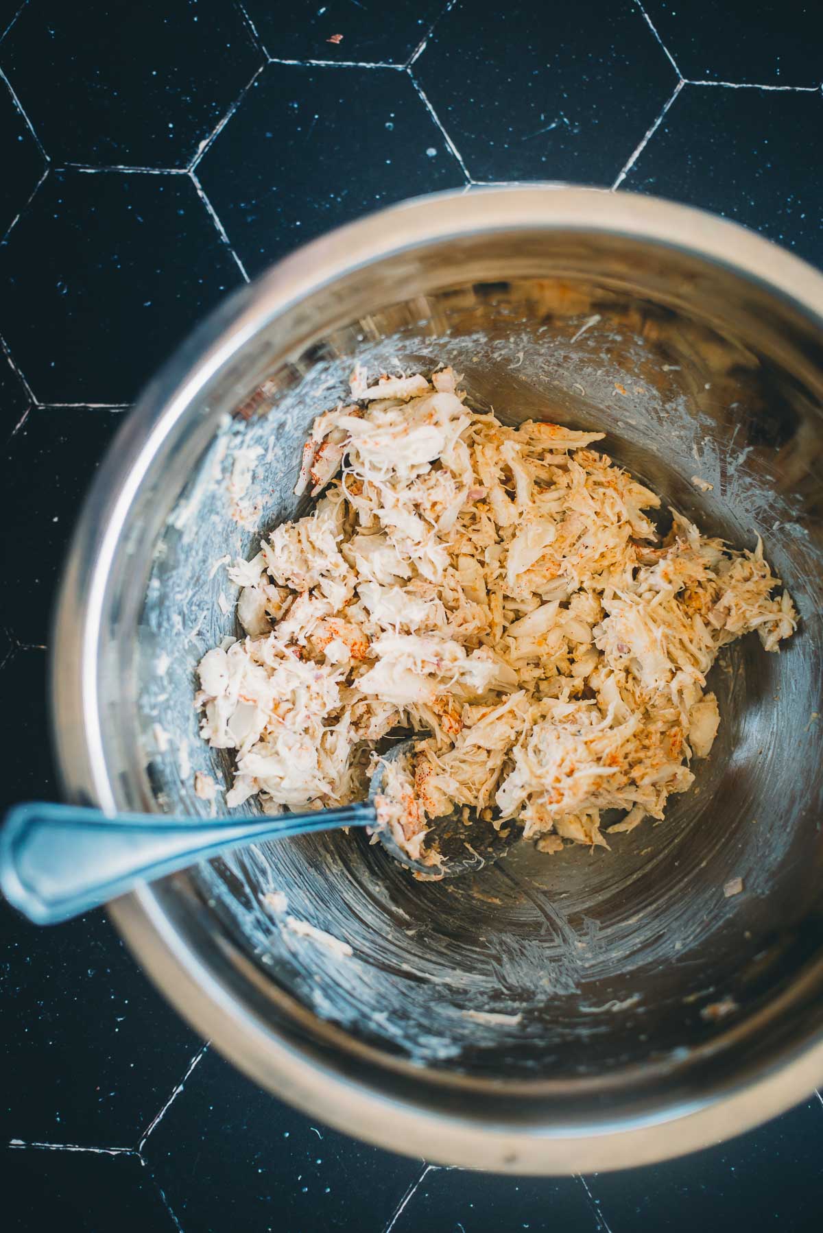 A metal bowl with crab meat and spices, placed on a black countertop. 