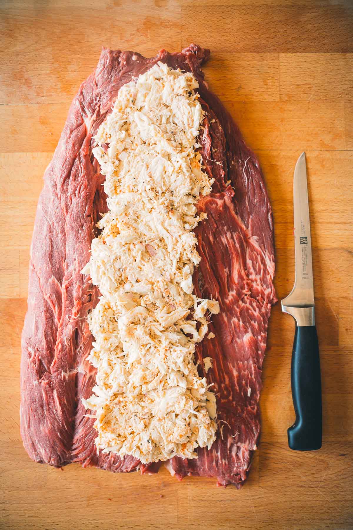 A butterflied beef tenderloin layered with crab filling on a wooden surface next to a knife.