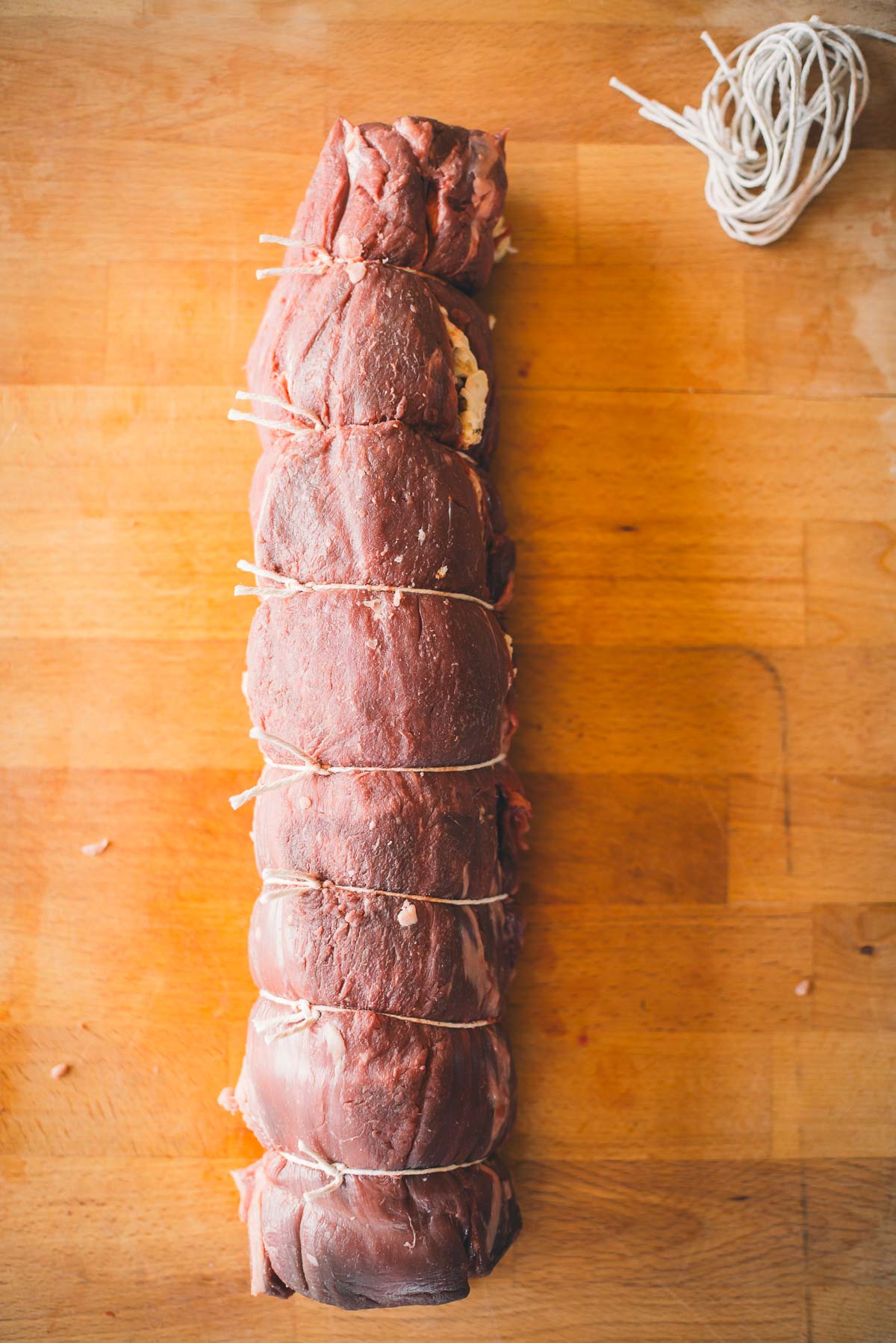 A tied, uncooked beef tenderloin roast on a wooden surface with a roll of twine nearby.