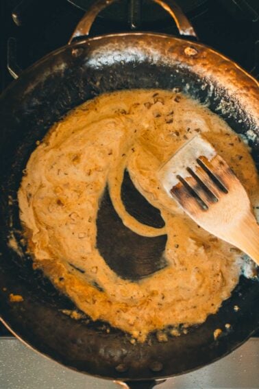 A wooden spatula rests on a pan containing a creamy mustard sauce with visible spices and herbs.