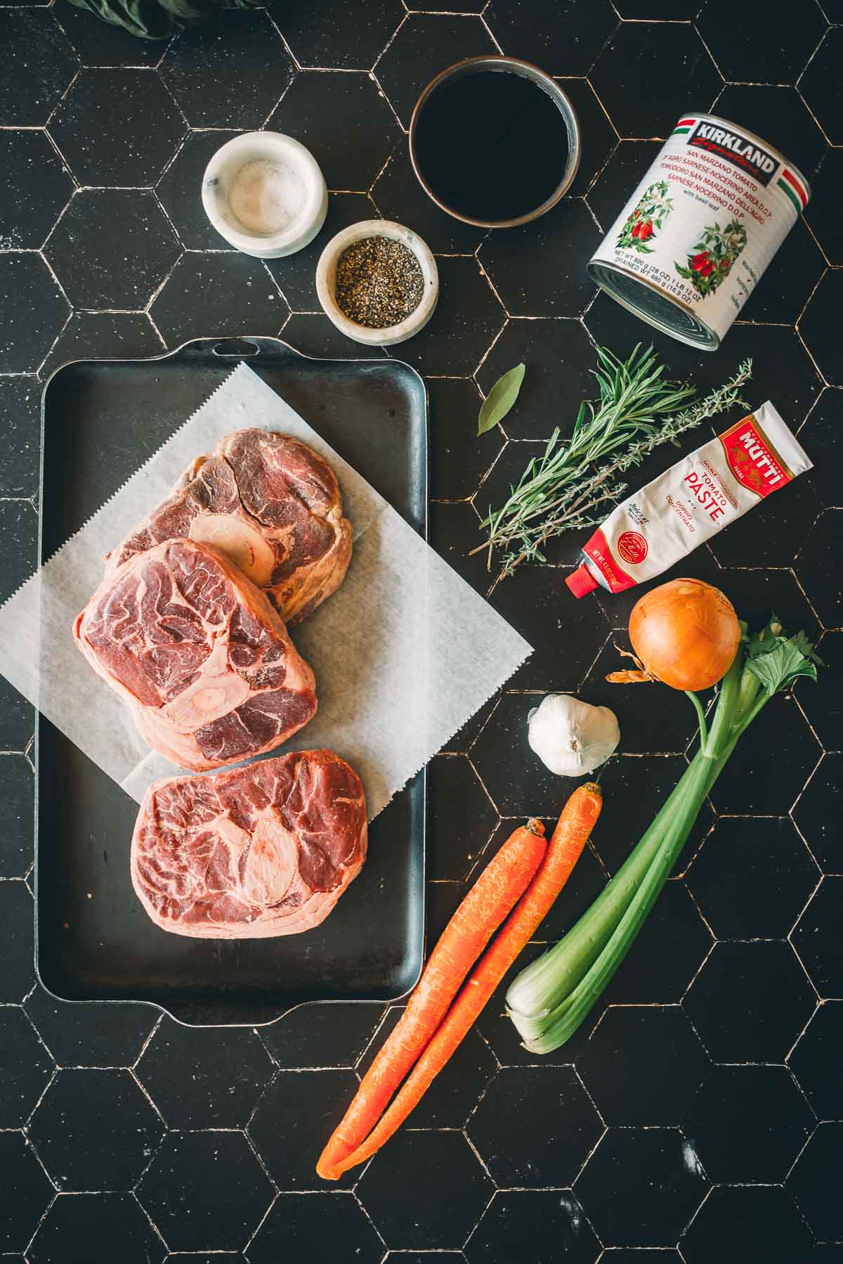 Ingredients for a slow cooker beef shanks: raw shanks on a tray, carrots, celery, garlic, onion, herbs, tomato paste, canned tomatoes, salt, pepper, and a bowl of sauce on a black tile surface.