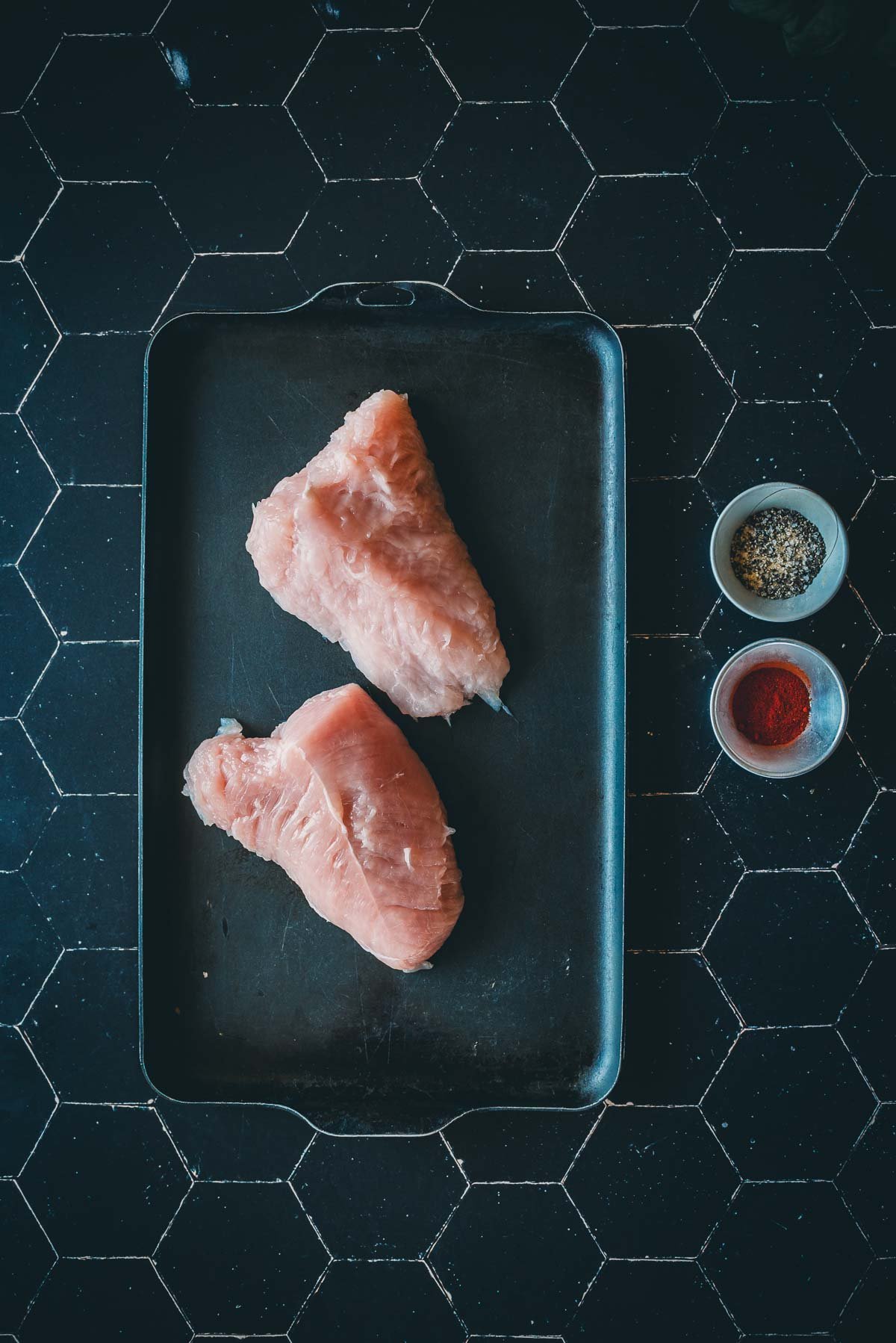 Two raw turkey breast tenderloins on a pan with small bowls of seasoning, including black pepper and paprika, on a dark tile surface.