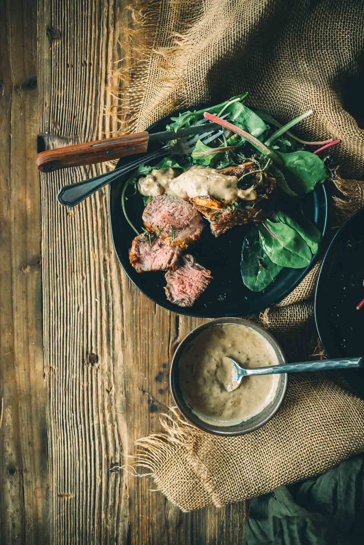 A plate of sliced wild boar chops and green salad with mustard cream sauce on a rustic wooden table, next to a small bowl with more sauce and a spoon.