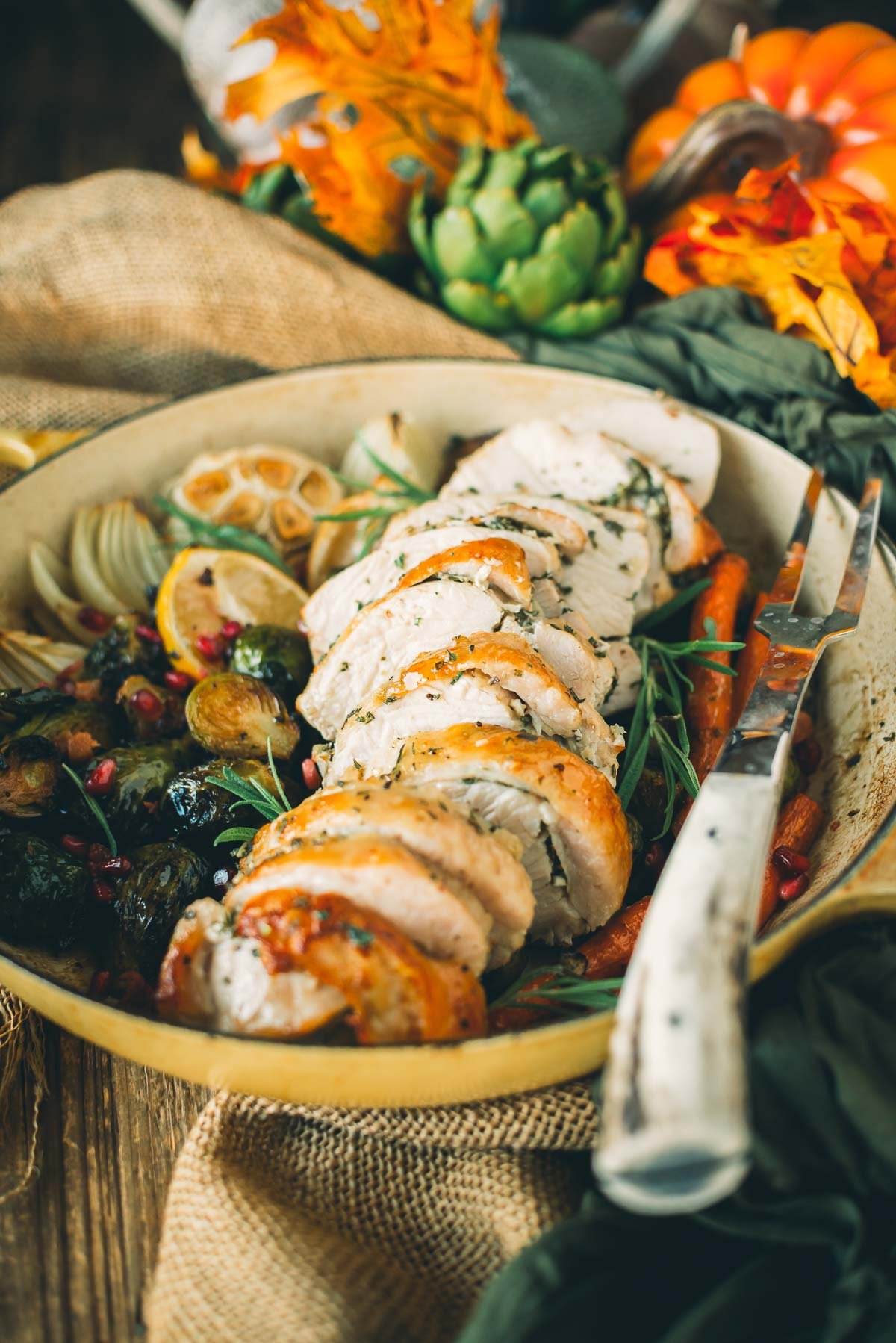 Sliced roasted turkey breast on a platter with Brussels sprouts, lemon slices, garlic, and garnished with greens; surrounded by autumn decor.