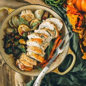 Sliced roasted boneless turkey breast with herbs in a pan, surrounded by vegetables like carrots, onions, and Brussels sprouts. Orange leaves and artichoke garnish the table setting.