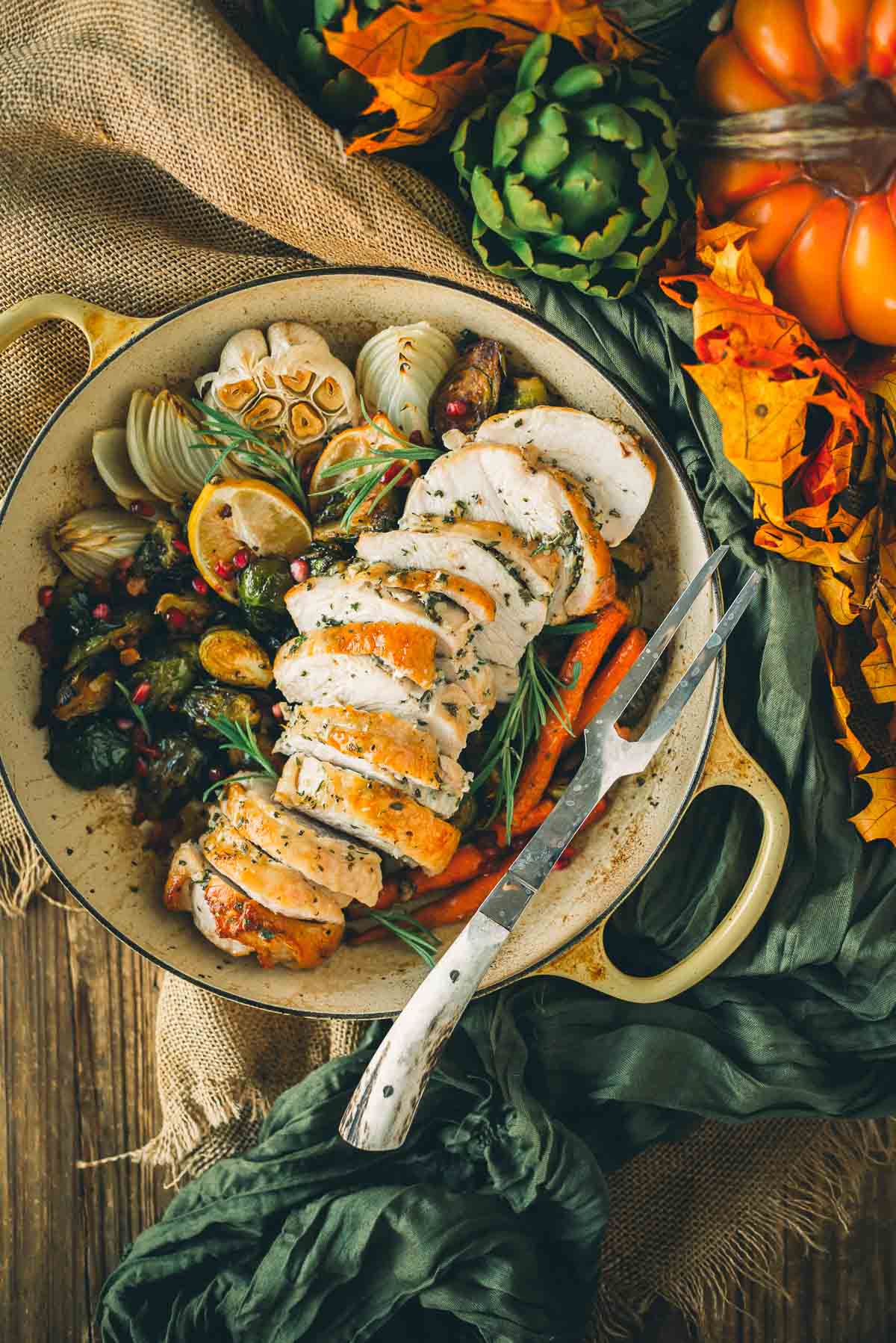 Sliced roasted boneless turkey breast with herbs in a pan, surrounded by vegetables like carrots, onions, and Brussels sprouts. Orange leaves and artichoke garnish the table setting.