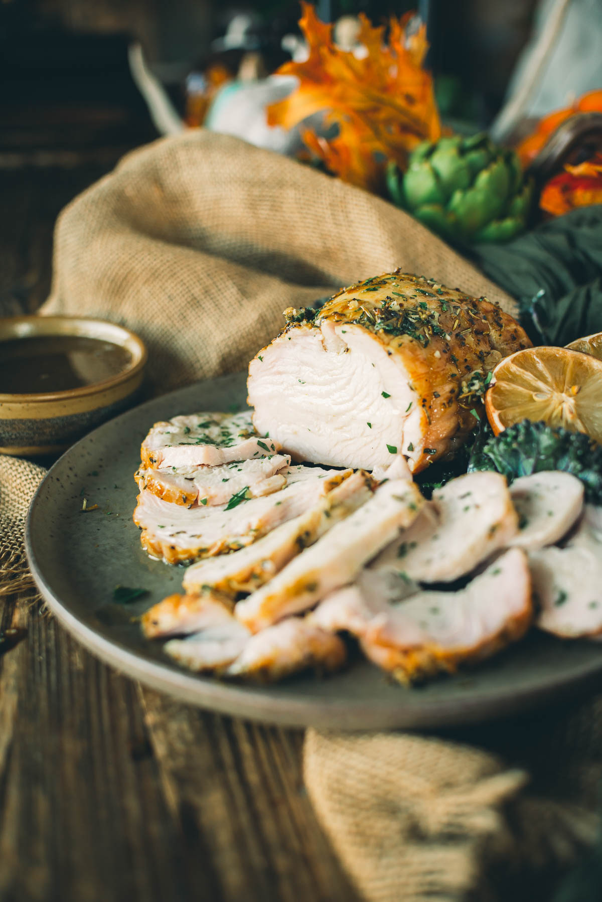 Sliced smoked boneless turkey breast garnished with herbs and lemon slices, served on a plate with a small bowl of gravy in the background. 