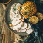 Plate of sliced boneless turkey breast with herbs, garnished with lemon slices, alongside dark leafy greens and a bowl of sauce.