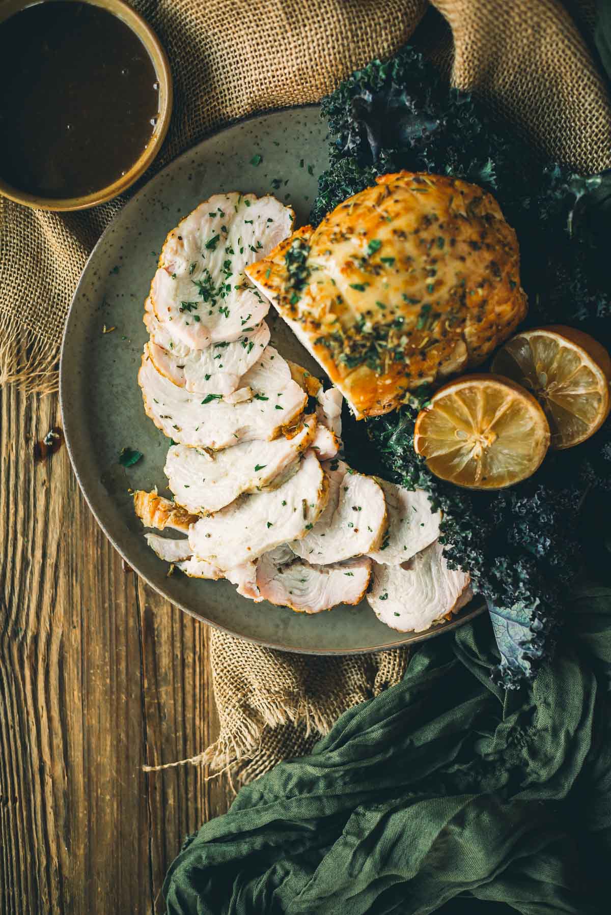 Plate of sliced boneless turkey breast with herbs, garnished with lemon slices, alongside dark leafy greens and a bowl of sauce.