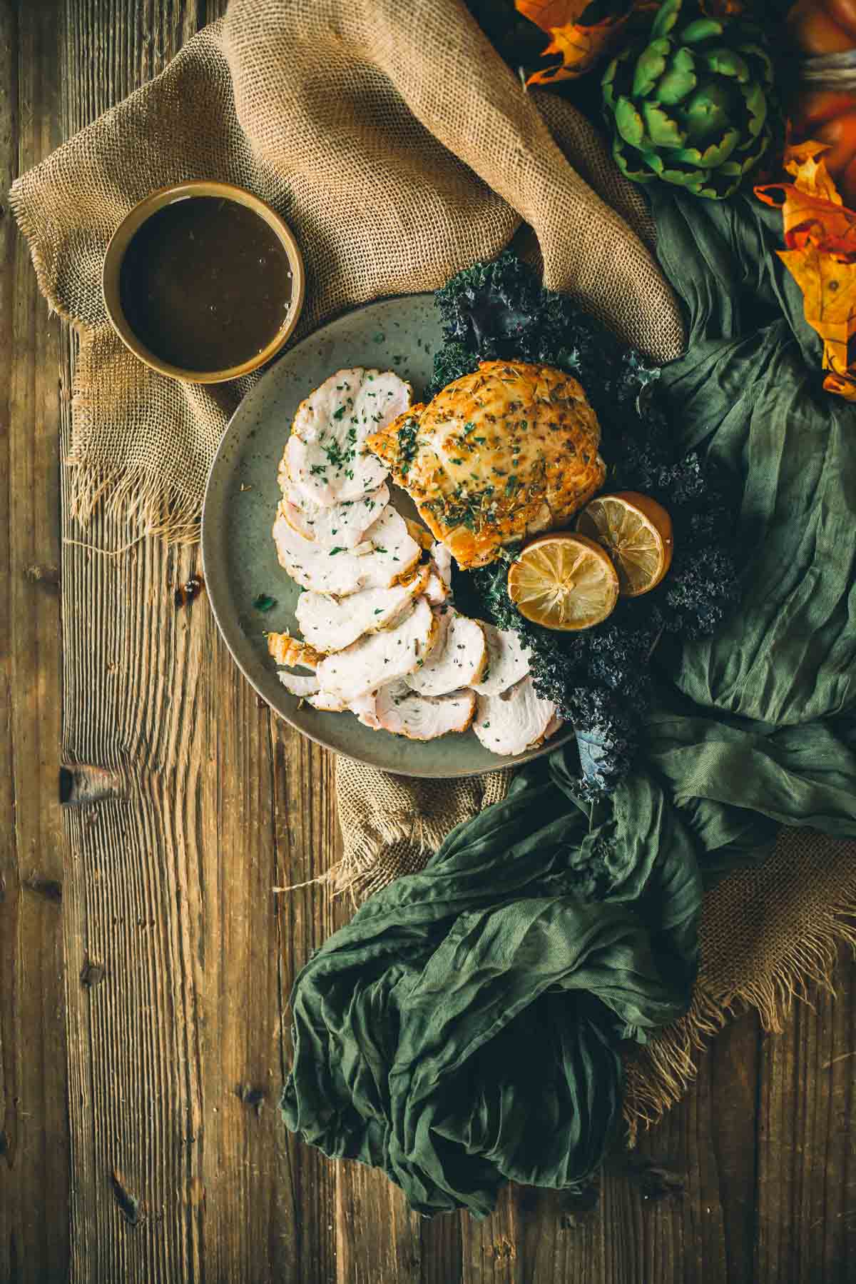 A plate of sliced smoked turkey breast with herbs, accompanied by kale, lemon slices, and a bowl of sauce, arranged on a wooden surface with burlap and dark green fabric.