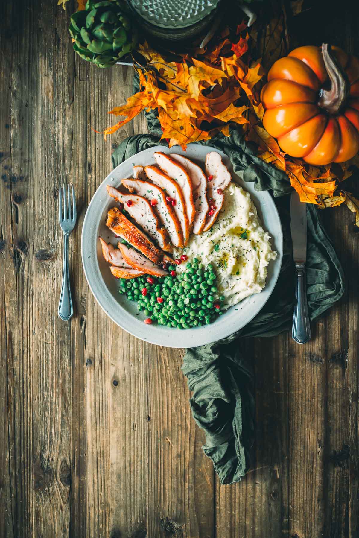 A plate of sliced smoked turkey tenderloin, mashed potatoes, and peas with pomegranate seeds is on a wooden table, surrounded by autumn leaves and a small pumpkin.