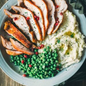 A plate with sliced smoked turkey breast tenderloin, mashed potatoes with gravy, and green peas, garnished with pomegranate seeds and herbs.