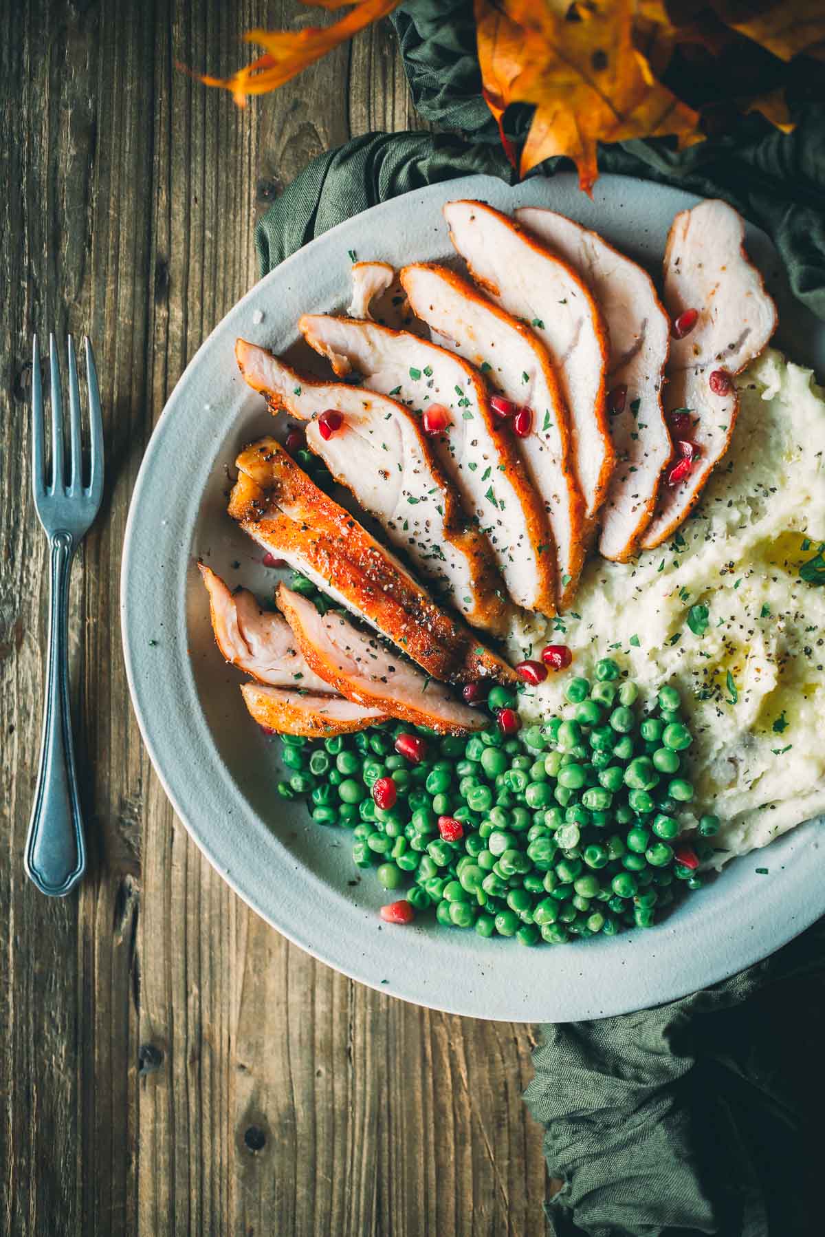 A plate with sliced turkey tenderloin, mashed potatoes, and peas, garnished with herbs and pomegranate seeds. A fork rests on a wooden table next to the plate.