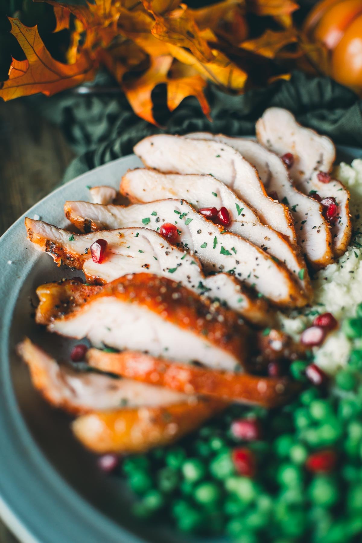 Sliced roasted turkey with garnish of herbs and pomegranate seeds, served with peas and mashed potatoes, on a plate with autumn leaves in the background.