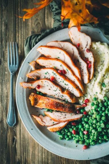 A plate with sliced smoked turkey breast tenderloin, mashed potatoes, and green peas, garnished with herbs and pomegranate seeds, beside a fork on a wooden table.