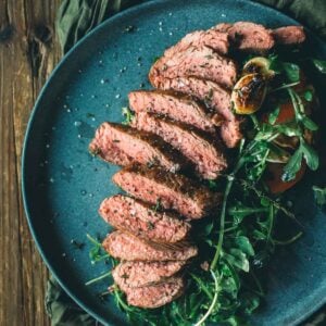 Sliced teres major steak on a blue plate with arugula and tomato garnish on a wooden table.