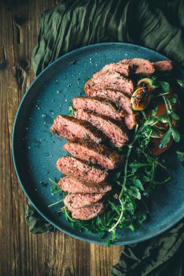 Sliced teres major steak on a blue plate with arugula and tomato garnish on a wooden table.