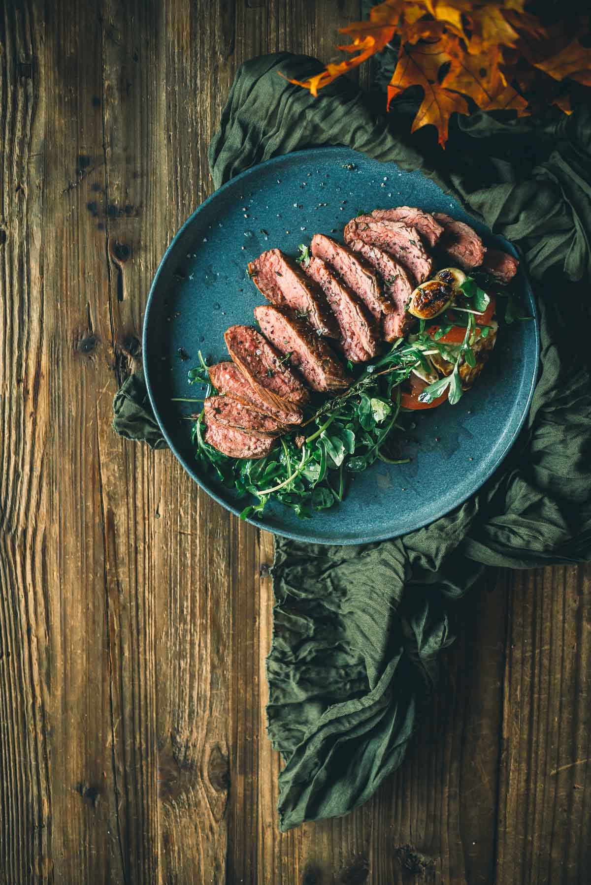 Sliced sous vide teres major steak on a blue plate, garnished with arugula and herbs, on a rustic wooden table with green fabric and autumn leaves.