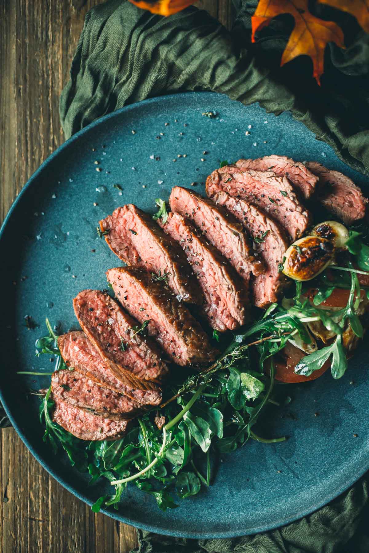 Sliced medium-rare teres major steak with arugula on a dark blue plate, garnished with a roasted garlic clove and sprinkled with salt.