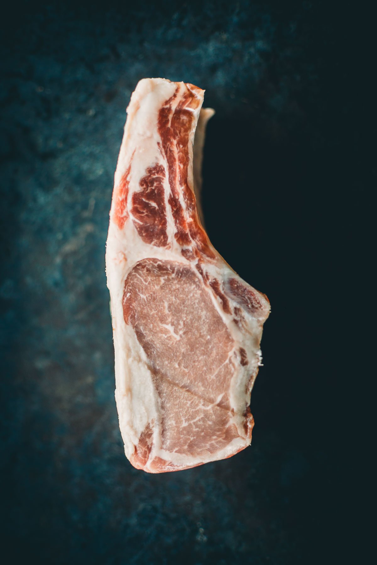 A raw, uncooked boar chop with bone, featuring marbled fat, against a dark background.