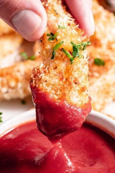 A hand dipping a crispy, breaded chicken tender into a bowl of red sauce, with more tenders and fresh parsley visible in the background.