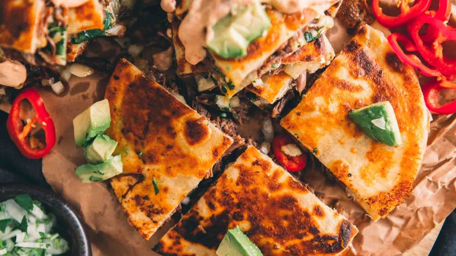 Close-up of grilled quesadillas with avocado chunks and sliced red chili peppers on top, arranged on parchment paper.