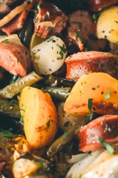 A close-up of a foil pack meal with sliced sausage, potatoes, green beans, and onions, garnished with herbs.