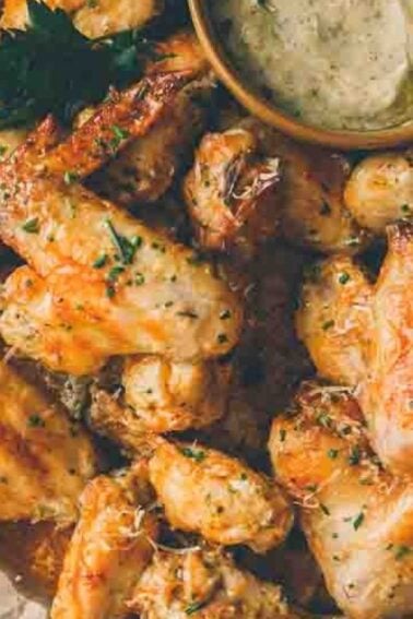 A pile of seasoned chicken wings garnished with herbs served on parchment paper, accompanied by a bowl of creamy dipping sauce.