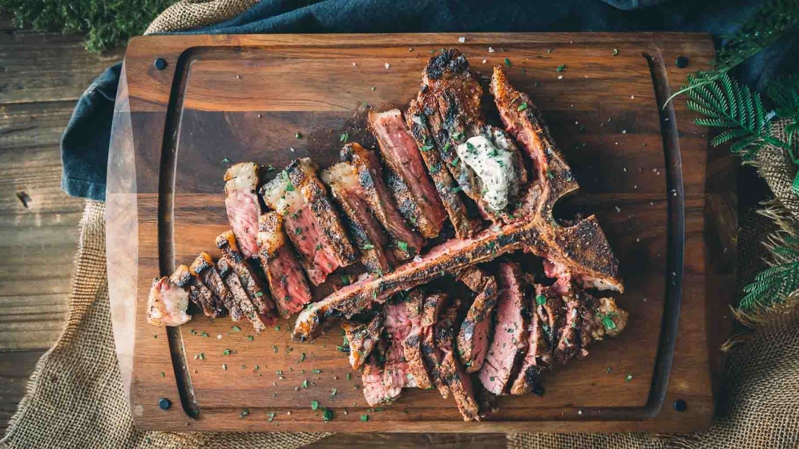 Sliced grilled steak with herbs and a dollop of butter on a wooden cutting board.