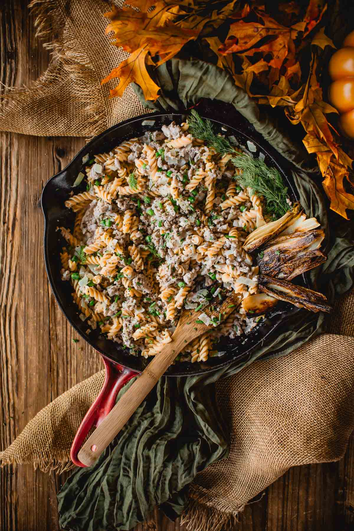 A skillet of ground turkey stroganoff, topped with herbs and grilled vegetables, on a rustic wooden table adorned with burlap and autumn leaves.