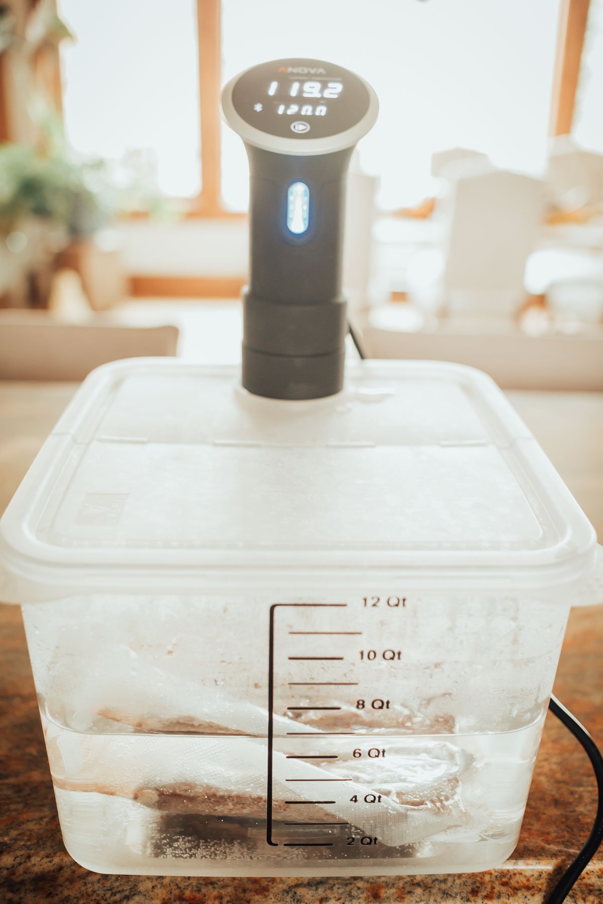 Sous vide machine with digital display showing temperature, attached to a water-filled container with steaks sealed in a plastic bag.