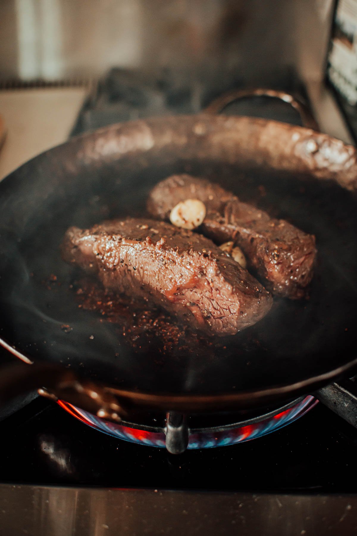 Two hanger steaks searing in a pan on a stovetop, with garlic slices visible on top.