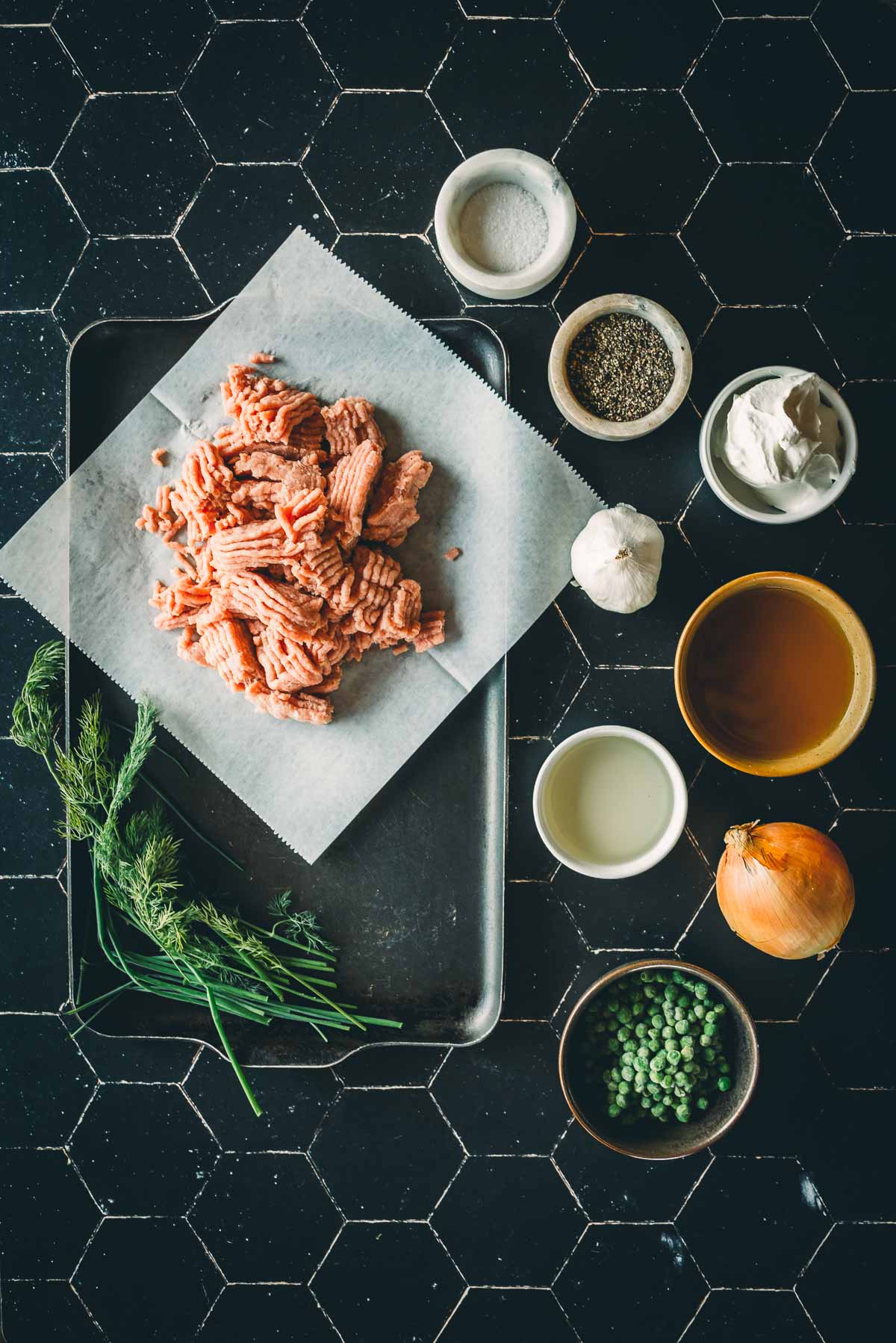 Ingredients on a tile surface: ground ground turkey on parchment, small bowls with spices, cream, and wine, a whole onion, garlic, fresh dill, and peas.