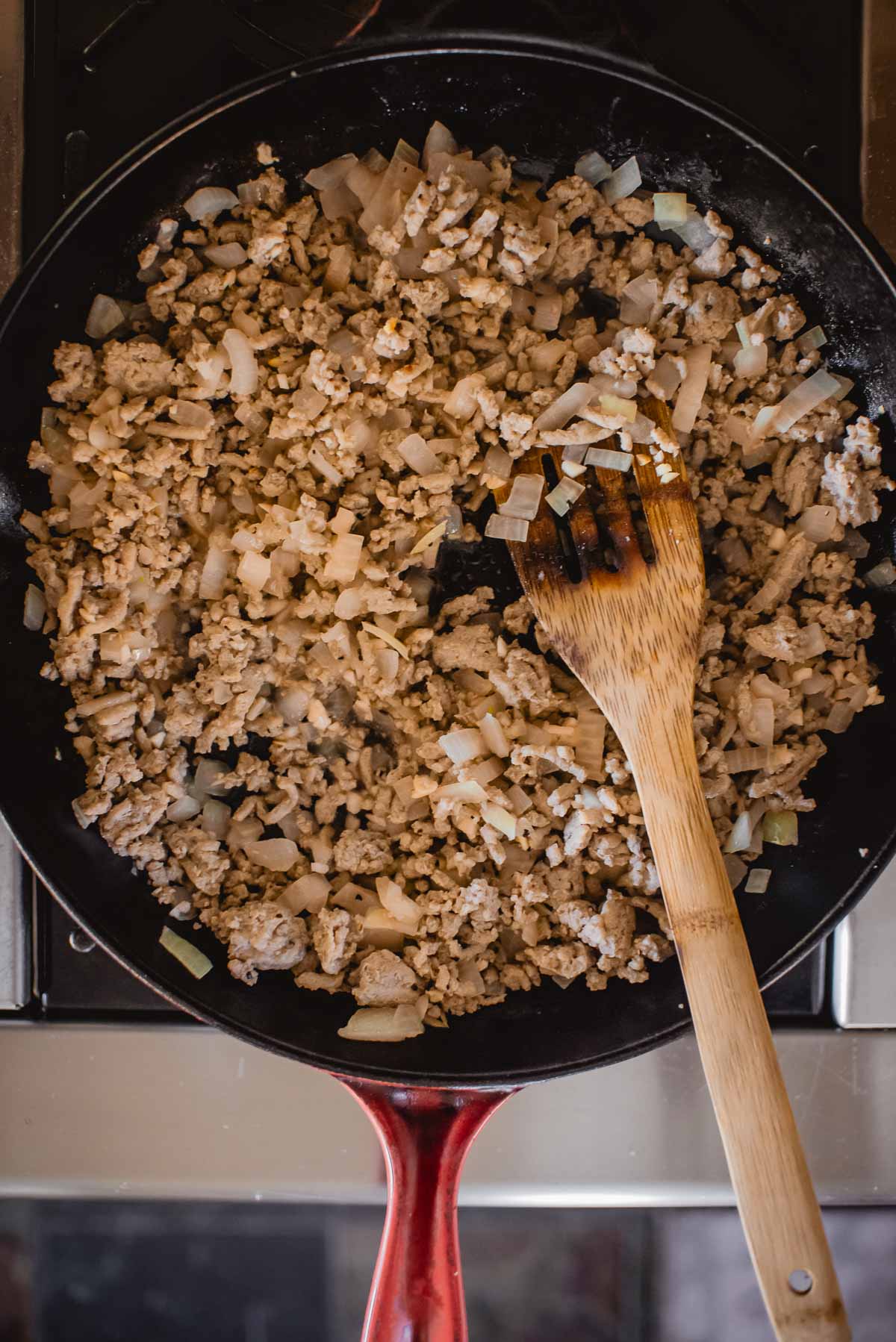 Ground turkey and chopped onions cooking in a red-handled pan with a wooden spatula.