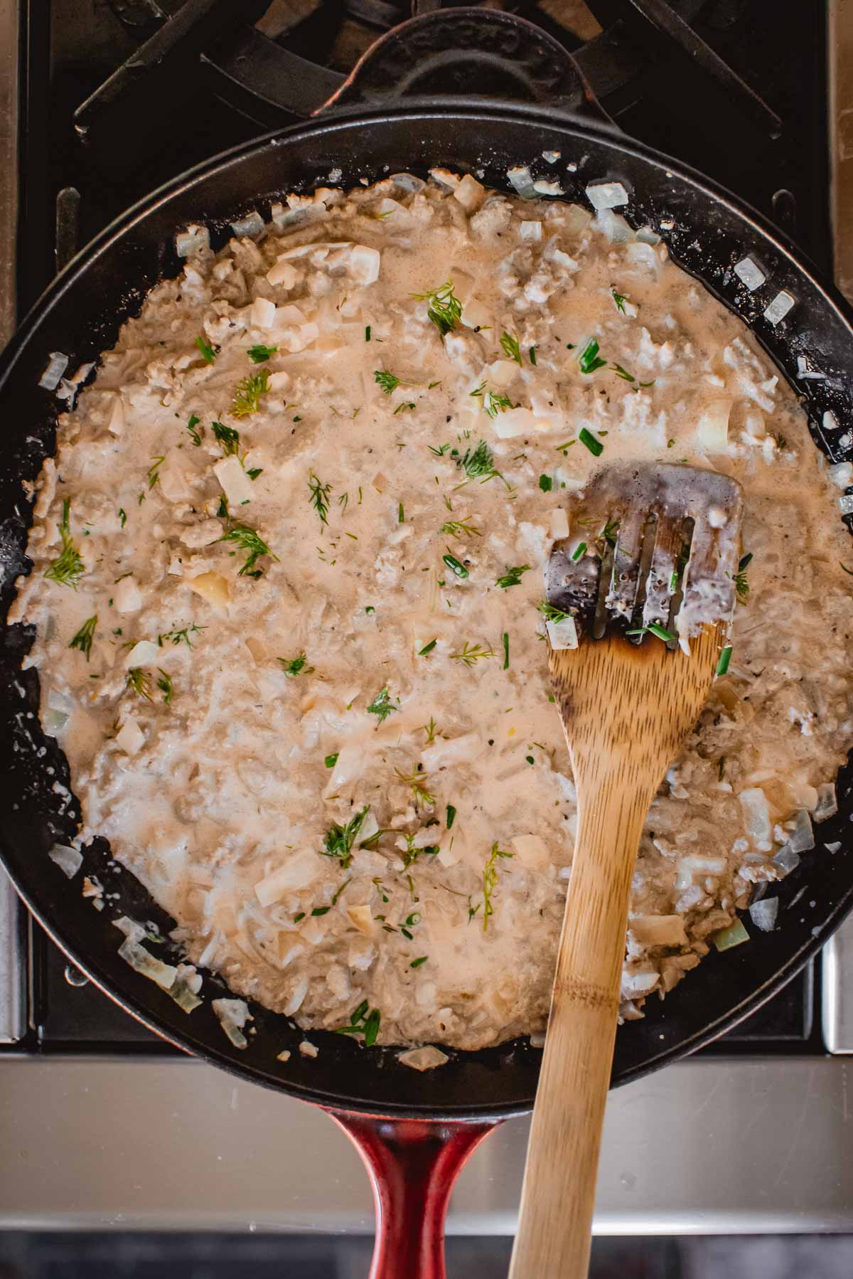 A skillet with creamy ground turkey and herb sauce, featuring chopped onions and herbs, with a wooden spatula resting on the edge.