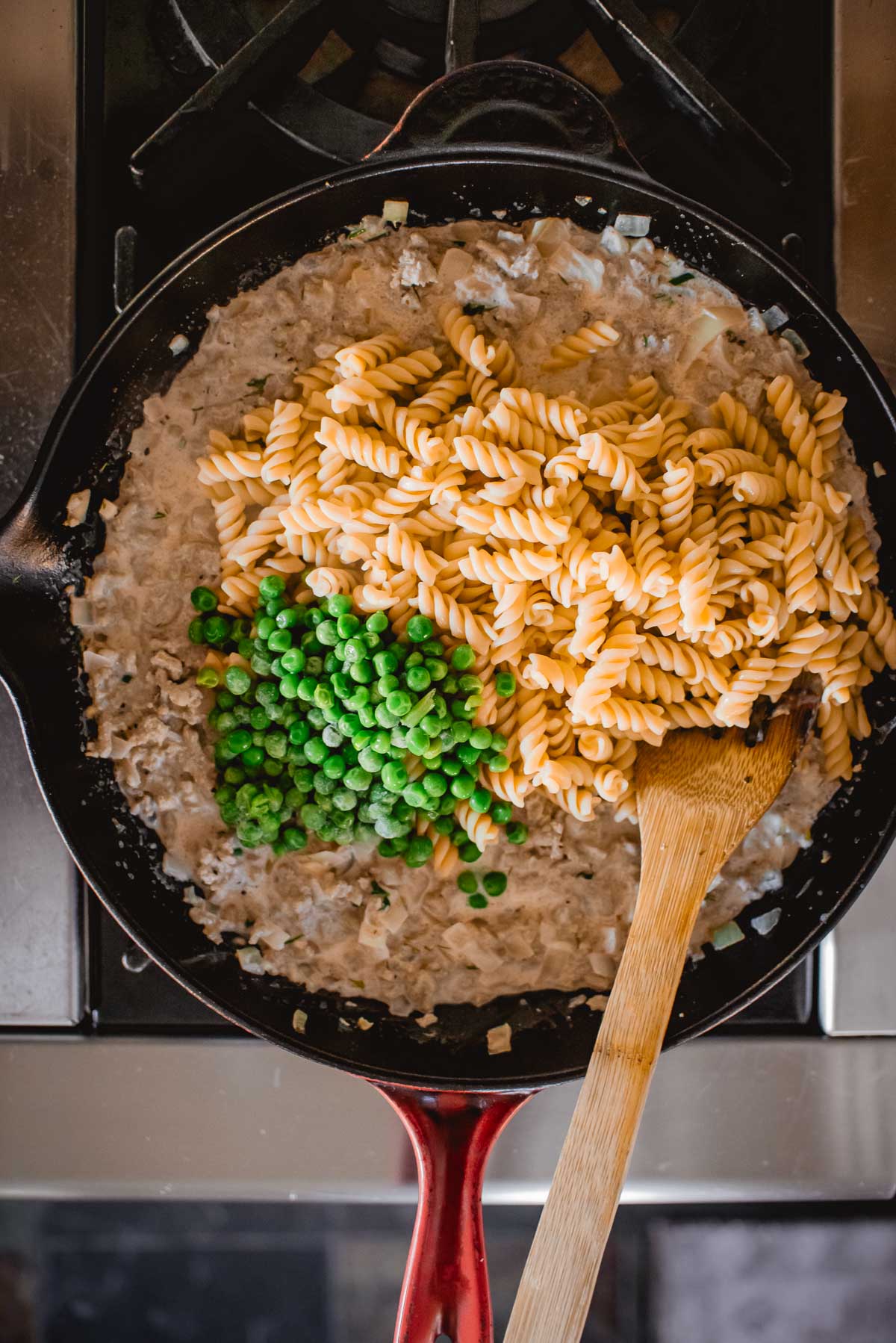 A skillet containing creamy sauce, pasta, ground turkey, and green peas, with a wooden spoon on a stove.