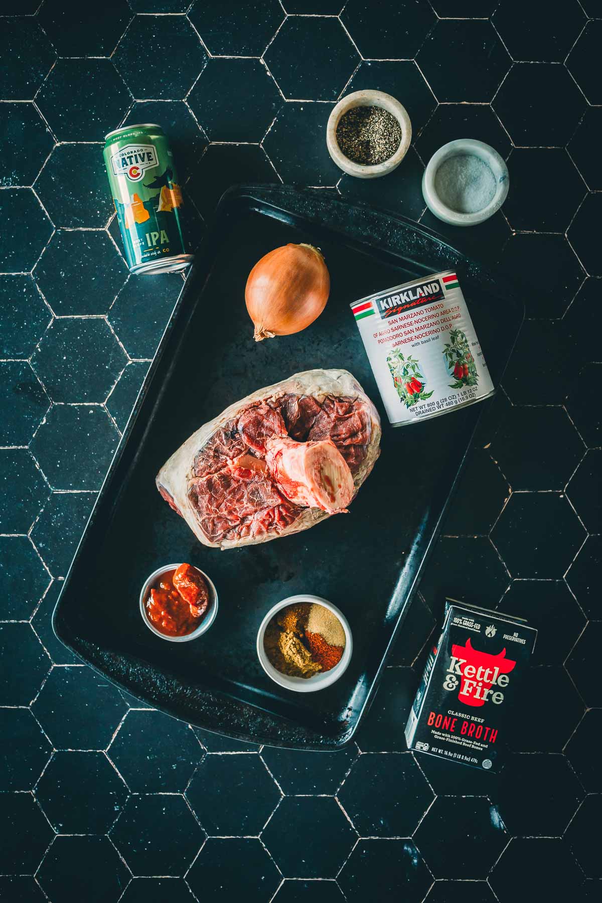 A tray with a beef shank, onion, and various seasonings, a can of beer, tomatoes, broth, and small bowls of spices on a black tile surface.