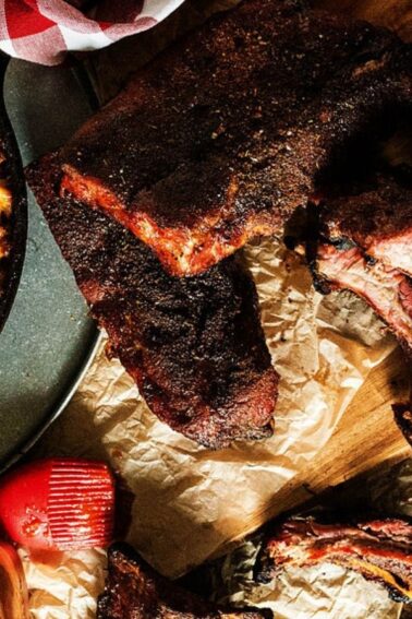 A dish of baked macaroni and cheese next to a cutting board with smoked barbecue ribs on a wooden table. A basting brush and a bowl of barbecue sauce are nearby.
