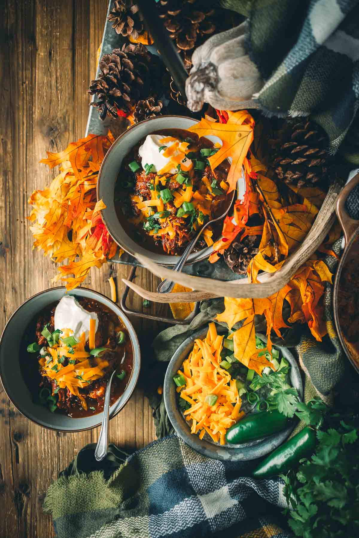 Two bowls of smoked over the top chili topped with sour cream and shredded cheese, surrounded by autumn leaves, pine cones, and a plaid blanket on a wooden surface.