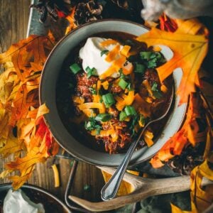 A bowl of smoked over the top chili topped with sour cream, cheese, and green onions, surrounded by autumn leaves and pine cones.