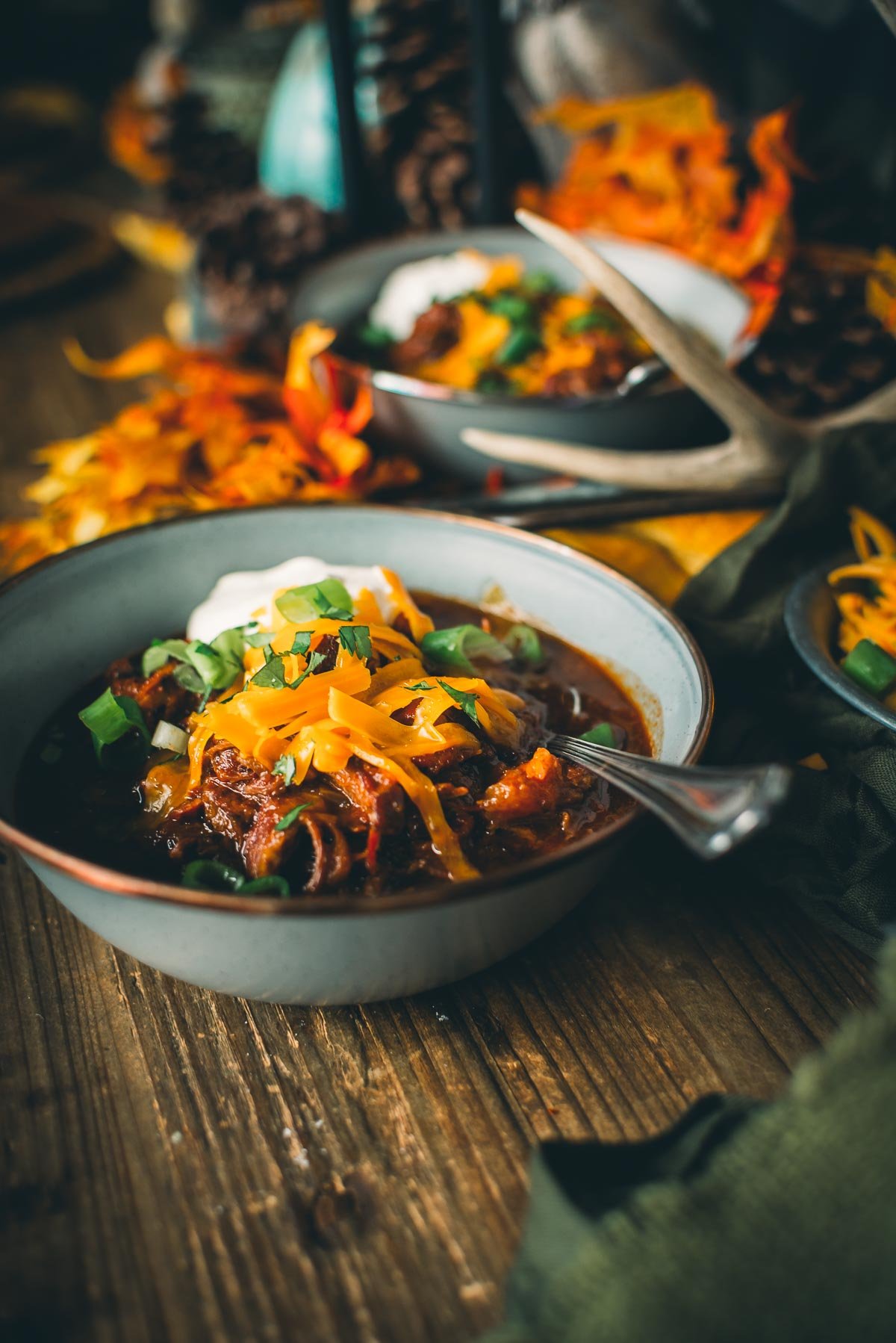 A bowl of over the top chili topped with shredded cheese, sour cream, and green onions, surrounded by autumn leaves and decor on a wooden table.